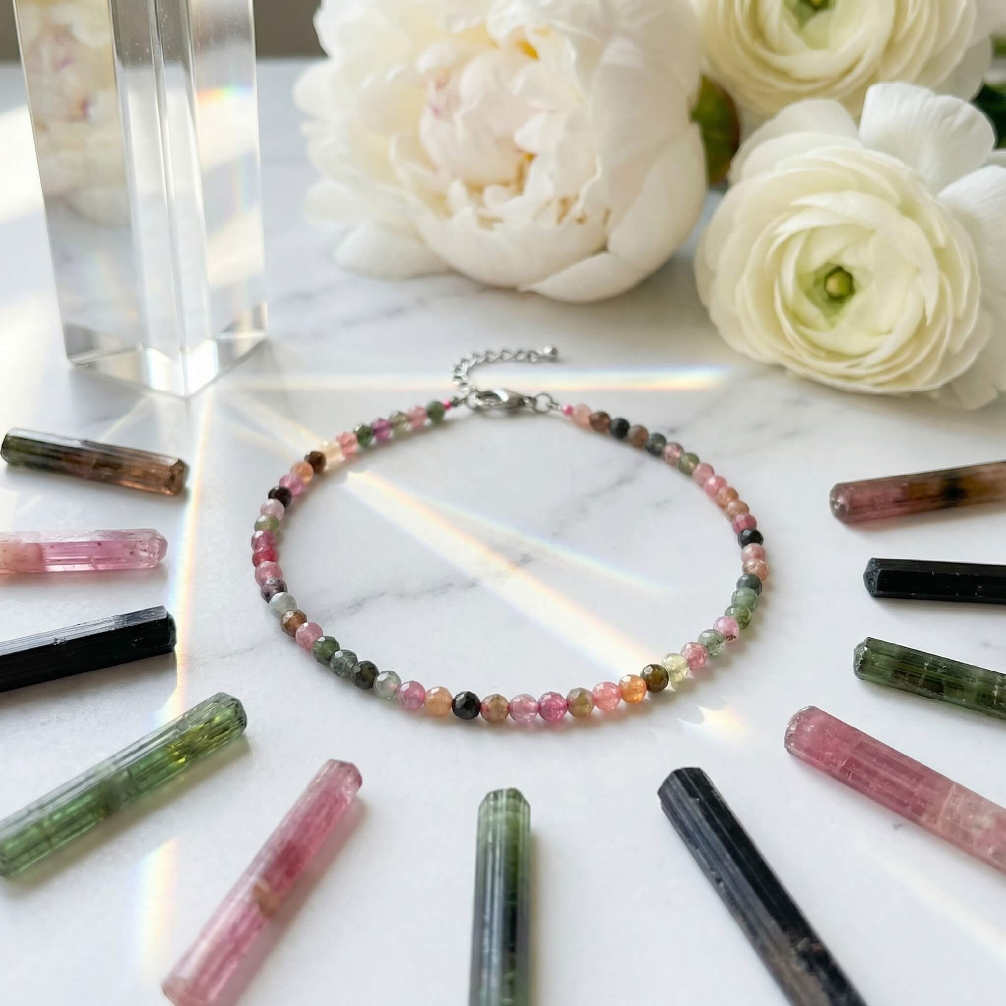 Multi tourmaline anklet on white surface surrounded by scattered multicolour crystal chips and white flower petals